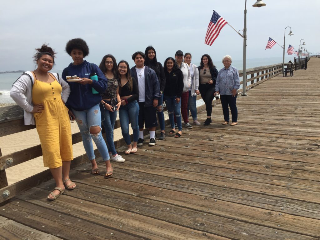 The group of people is standing on the wooden bridge near the ocean