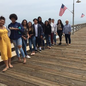 The group of people is standing on the wooden bridge near the ocean